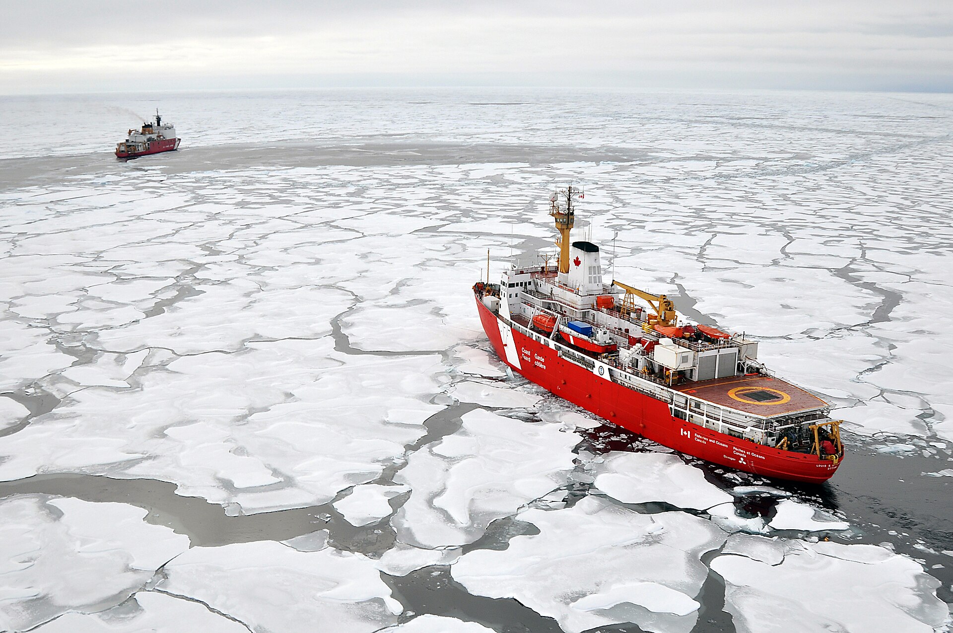 A red-and-white Canadian Coast Guard icebreaker pushing through white Arctic pack ice under a grey sky